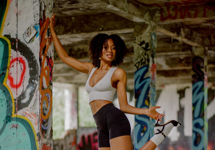 Writer Racheal Ofori poses, leaning against a concrete pillar while holding onto her heeled black shoe. 
