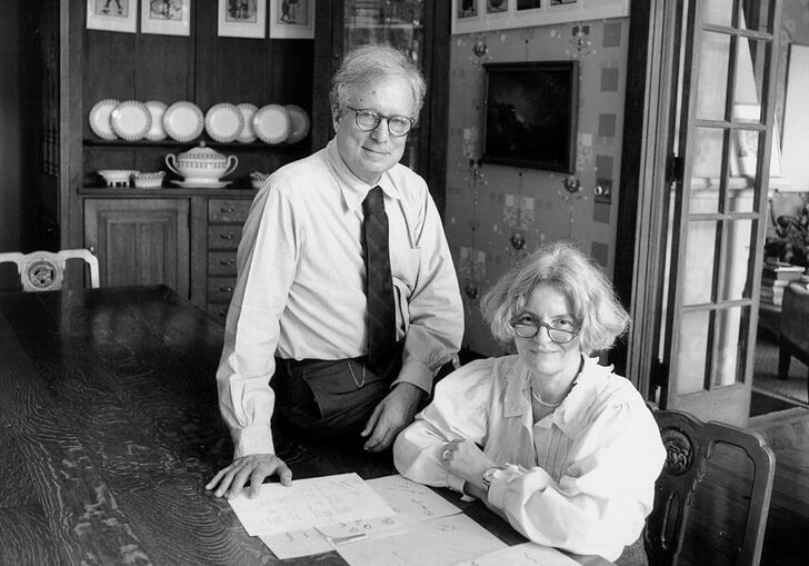 Black and white photo of Robert Venturi and Denise Scott Brown. She is sat at a large table and he is next to her, perched on the table.