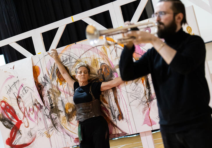 Rachel, a white woman with short cropped hair, wearing black, and Freddie, a white male, with a full beard, wearing a black long sleeved top with a black skirt, in rehearsals for PostHuman. Freddie is out of focus in the foreground playing a silver trumpet, and Rachel is in focus leaning back on a large canva with arms out and eyes closed.