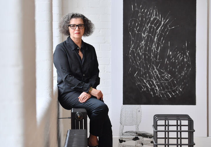 Mona Hatoum sitting on a radiator on the wall of her studio. On the wall next to her is Untitled (Bed Springs), one of her lithograph print depicting a structure made of bed springs, curved and crumpled.