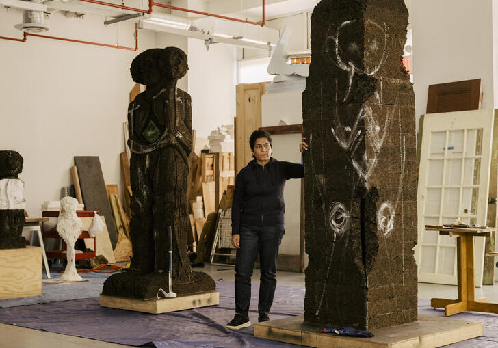 Huma Bhabha standing in a studio with two tall sculptures, one carved out and one with markings ready to be carved.