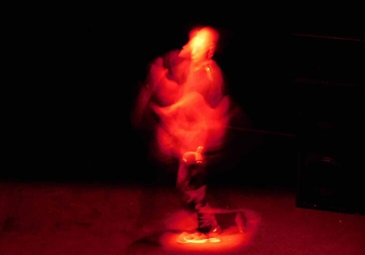 A person moves through a dark space, they are lit by a red light so that you can't see them as much as just a red figure moving through the space.