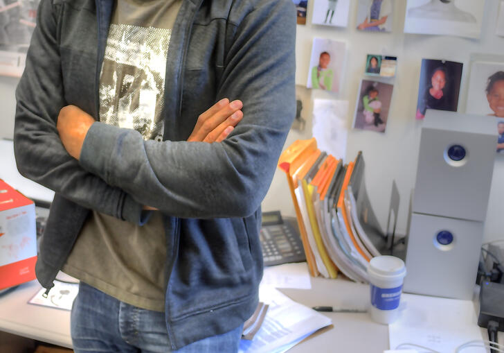 A man stands leaning against a desk. The wall behind is filled with images.