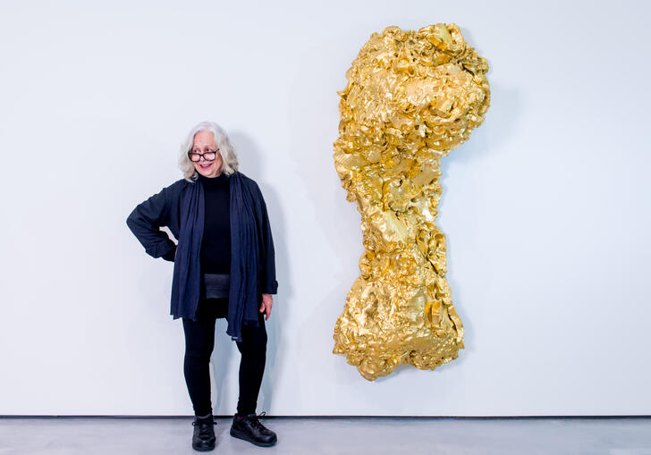 Lynda Benglis standing next to one her sculpture titled Ghost Dance/Pedmarks. It is a large, gold, torso-like form hanging on a white wall.