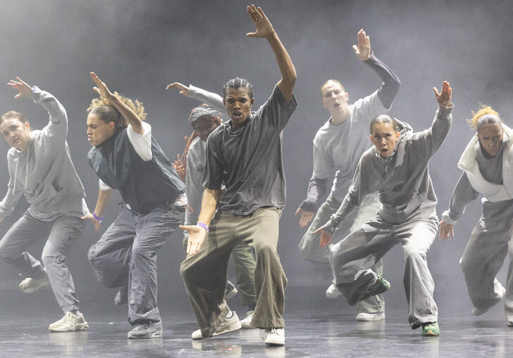 A group of hip hop dancers caught in motion. They lift one arm in the air and open their mouths.