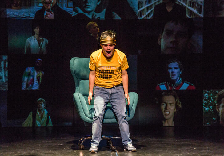 A person in a yellow shirt and blue jeans standing over a green sofa chair, shouting. They are against a background of screens with each screen showing pictures of different men.