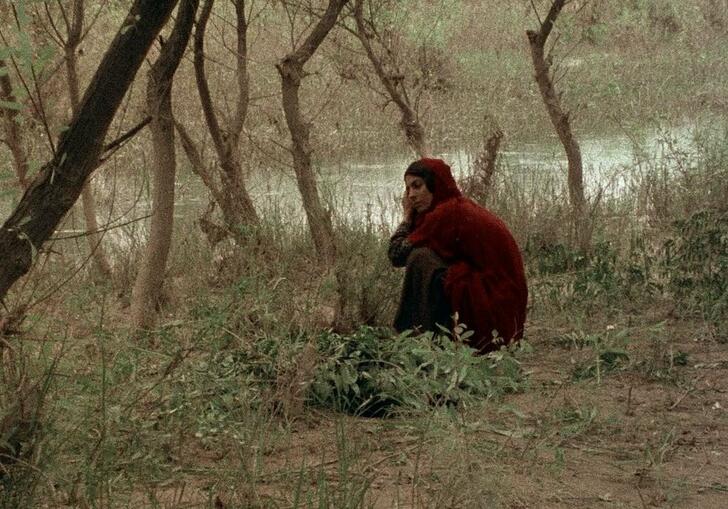 A woman wearing red, crouching amongst the trees by a river.