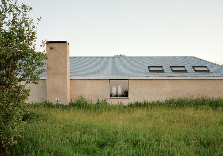A photo of the Farmworker's House in Cornwall. It is a single storey, with a lime render and a pitched roof with three roof windows on the right side. On the left is a large chimney. It is surrounded by a large meadow of grass.