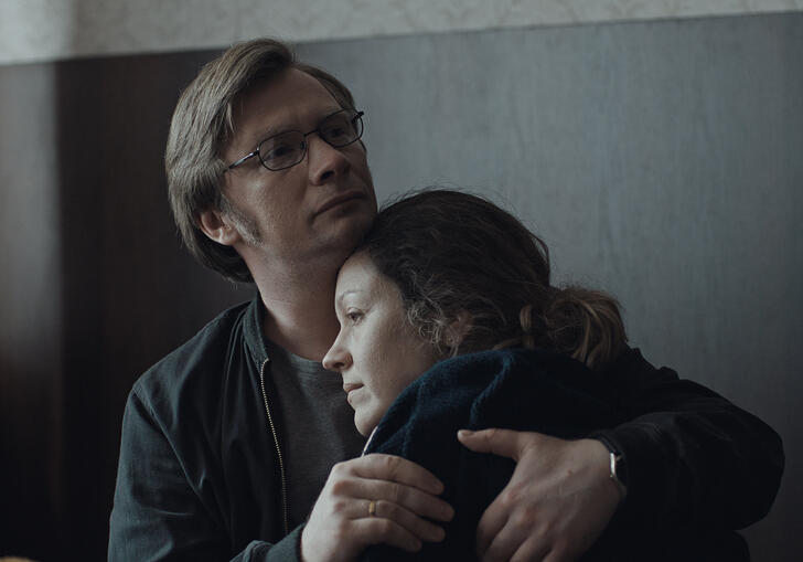 A man and woman hug in a grey room. 