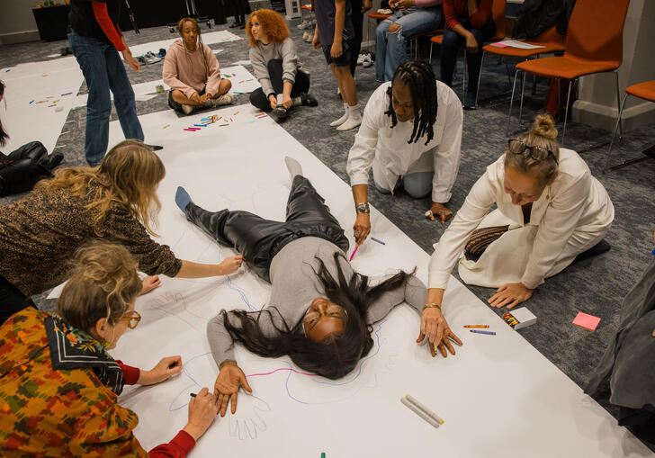 A person lies on the floor on a large piece of white paper. A group of people draw around them using a range of pens and pencils. 