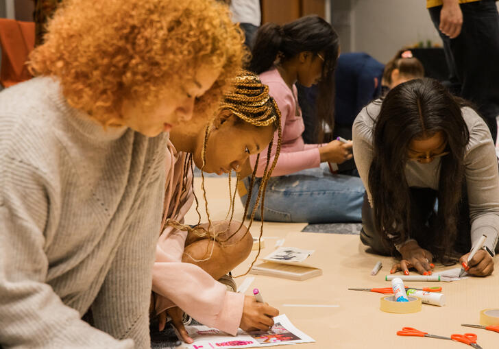 A group of people sit beside each other on the floor, drawing, colouring and collaging.