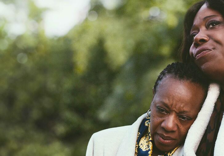 Two women in a park - one resting her head on the shoulder of the other.
