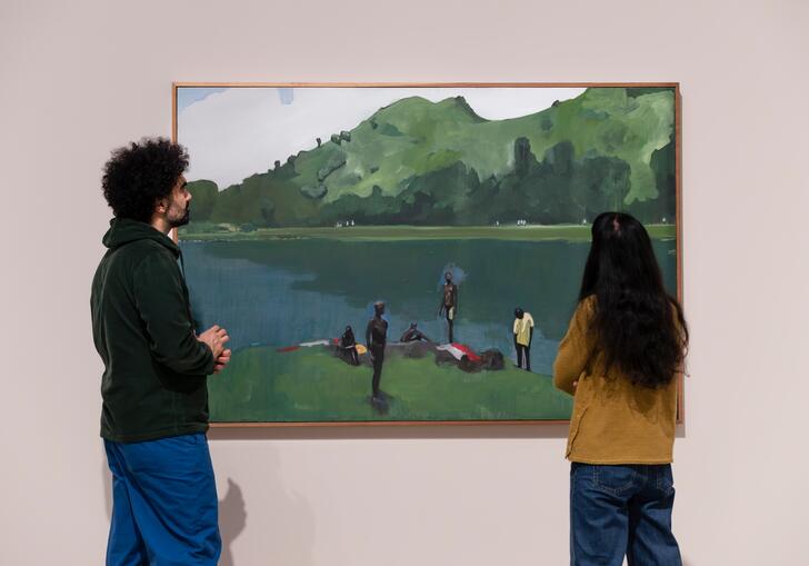 A man and a woman stand in front of a large painting depicting people lounging on the grass next to a large body of water.