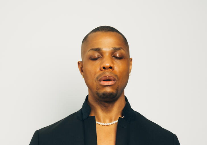 Davone Tines wearing a black suit with his eyes closed and hands in front of his chest