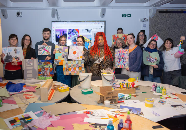 A group of people hold their artwork up proudly in front of a table filled with craft objects.