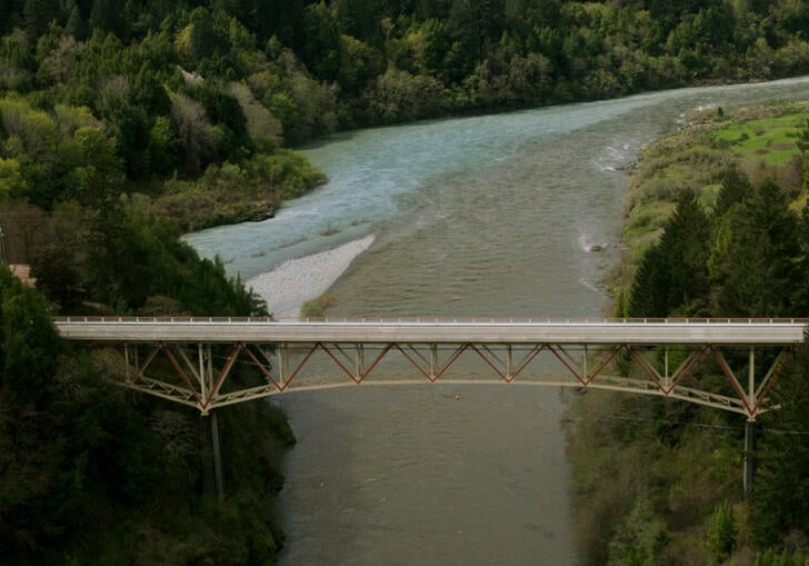 A still from Lucy Raven's moving image installation called Murderers Bar. It shows a bridge over a large river, surrounded by tree-covered hills.