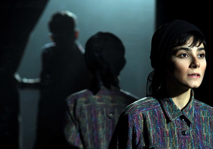 A woman is lit in the foreground is reflected by a mirror, as someone else in shadow walks through a curtain. 