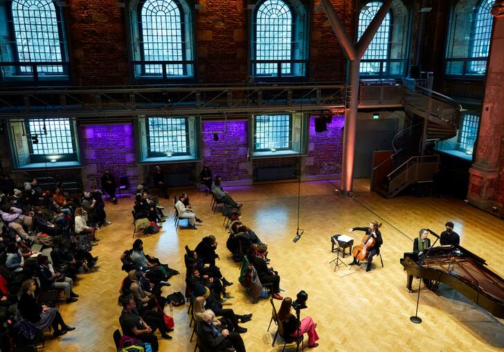 A view of the Jerwood Hall in LSO St Luke's from the balcony, looking down on rows of audience members sitting on chairs, facing a cellist and pianist on stage