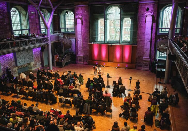 A view from the balcony at the back of the Jerwood Hall in LSO St Luke's, showing the audience sat in chairs and two musicians bowing on-stage