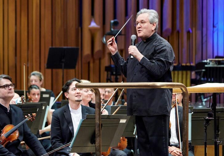 Sir Antonio Pappano standing on the podium facing the audience, talking to the audience, with the London Symphony Orchestra sat on-stage behind him
