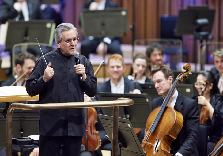 Sir Antonio Pappano standing on the podium on the Barbican stage, facing the audience and talking into a microphone