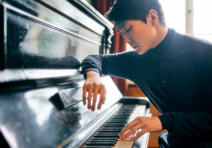 Seong-Jin Cho sat at a piano, facing towards the keys, with his arm resting on the lid of the instrument