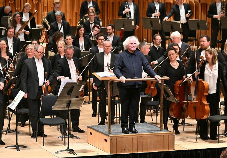 Sir Simon Rattle standing on the podium on the Barbican stage, facing the audience and smiling, with the London Symphony Orchestra standing behind him