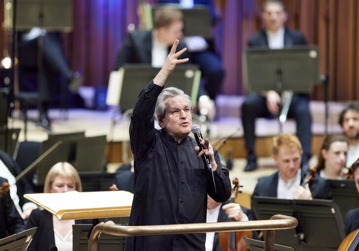 Sir Antonio Pappano standing on the conductor's podium, facing the audience, talking into a microphone with his right-hand raised