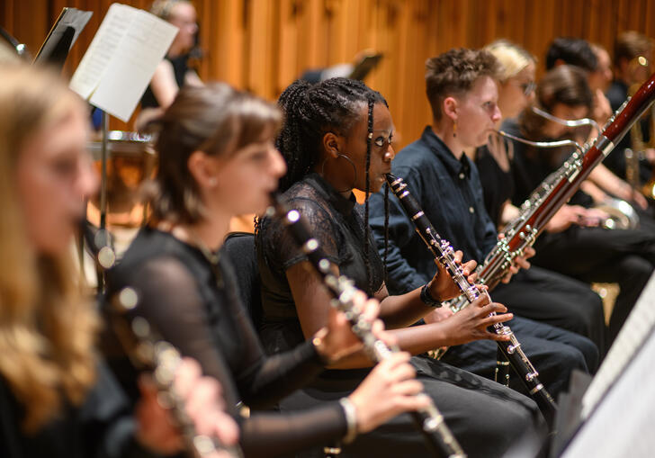 Image of an orchestra performing with multiple people stood in line playing the flute