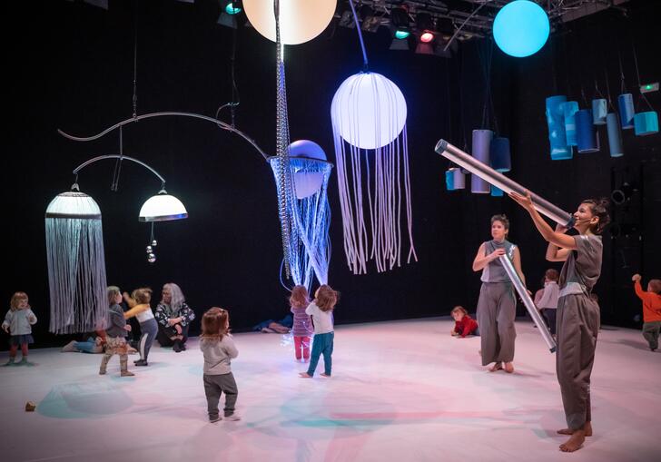 Young children run around a space interacting with large, colourful objects in a sensory show.