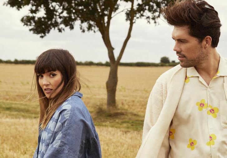 An image of both members of Oh Wonder walking away from each other in a field of grass with a tree behind them in the distance