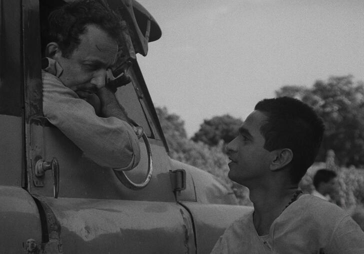 A black and white still from Badnam Basti, showing a man leaning out of a truck window speaking to another man.
