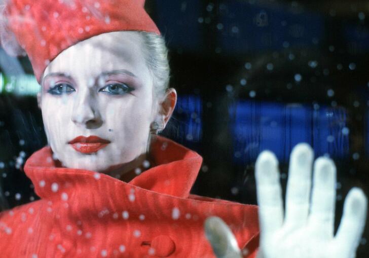 A woman in a red coat and red hat stands in front of a window with her hand placed up on the glass.