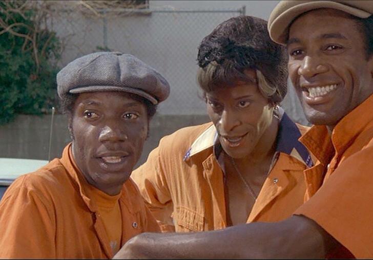 Three people in orange overalls chat together and stare into the camera
