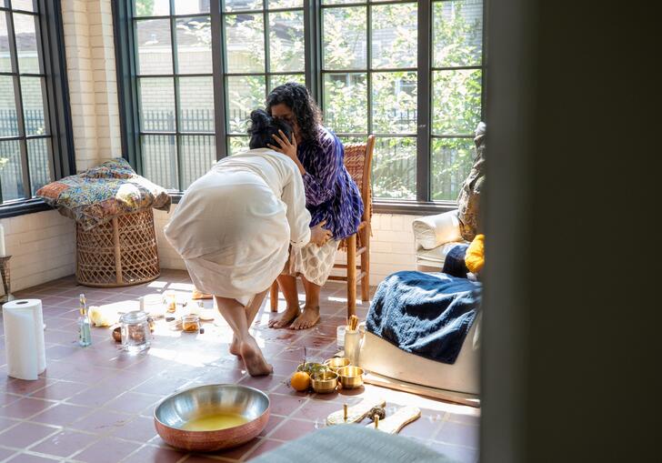 A woman crouches in front of another woman who is kissing her forehead, showcasing a moment of care and companionship.