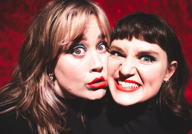 Two people wearing black against a red background, smiling with cheeks pressed together