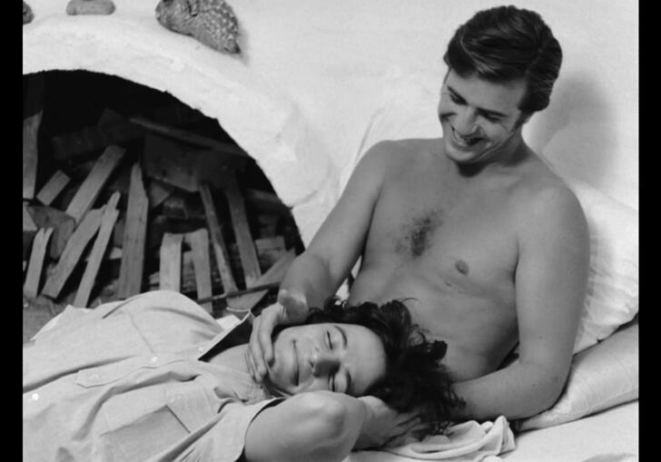 A black and white still from The Set, showing a topless man laying in bed with another man laying in his lap.