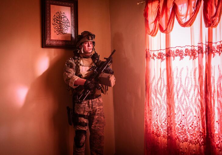 An American soldier stands in a room flooded with pink light, hiding in the corner next to the window. 