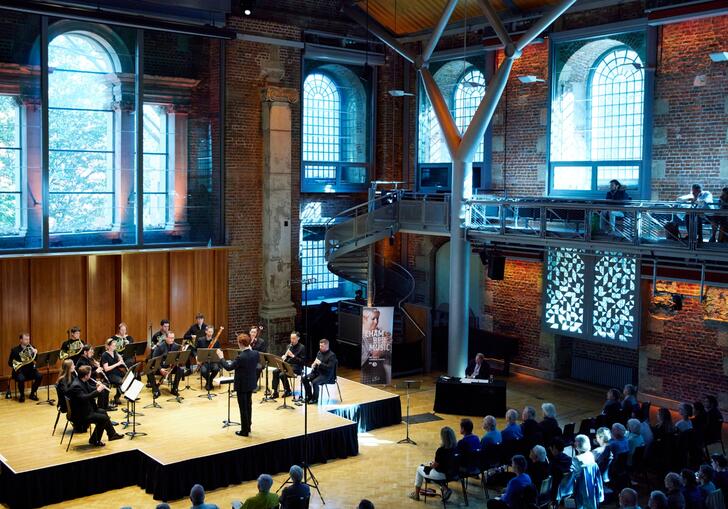 A view of the Jerwood Hall in LSO St LUke's from the balcony, with a chamber group of LSO musicians sat performing on the stage, an audience watching, and sun coming through the venue windows