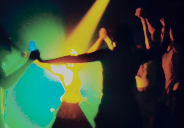 Figures are dancing in a dark room lit by yellow, green and blue lights.
