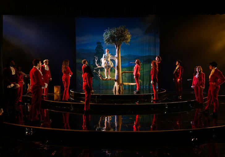 Orlando stands on a tree prop center stage, surrounded by a semi-circle of actors in matching red suits against a painted countryside backdrop."