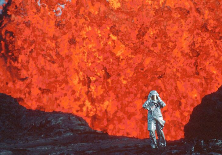 A person in a protective suit standing in front of an erupting volcano.