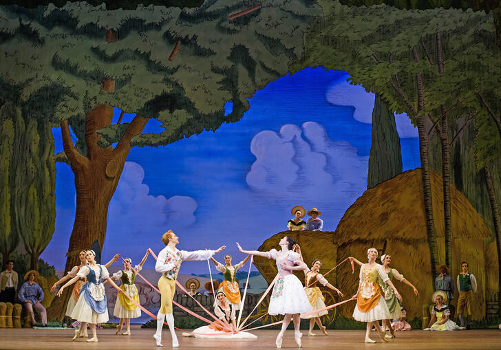 Ballet dancers on stage with ribbons, set against a countryside painted background