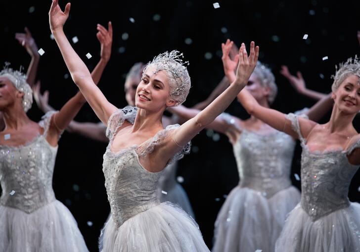 Ballet dancers dressed as snowflakes dance with their arms raised, snow falling around them