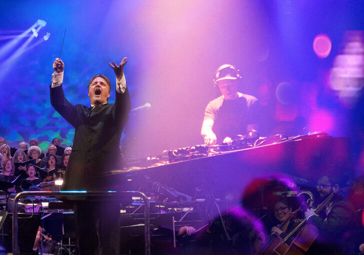 Conductor Fernando Furones leads an orchestra in the foreground with an image of a choir on the left hand side and an image of Symphonic standing behind a DJ deck on the right hand side