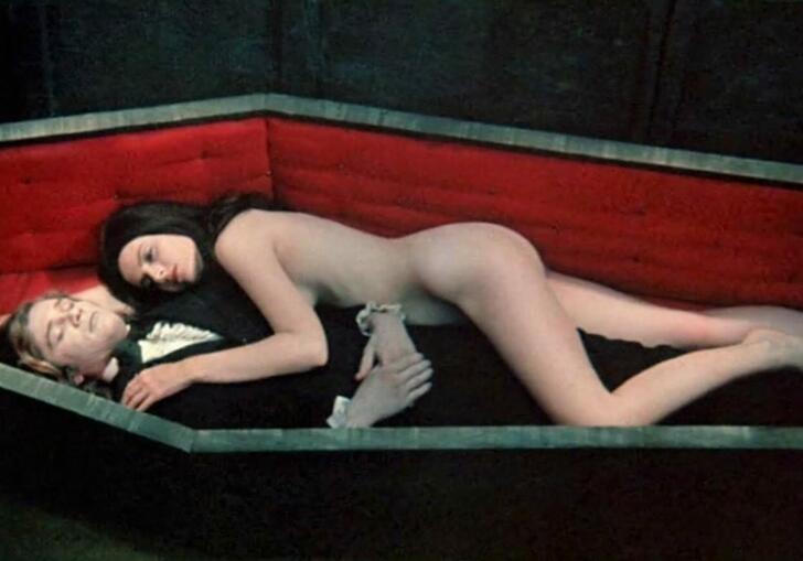 A naked woman lies on a man in a suit in a coffin.