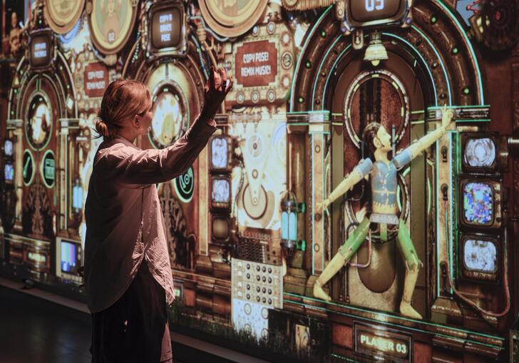 A woman stands in front of a large wall projection, mimicking the dance move shown by the character on the screen