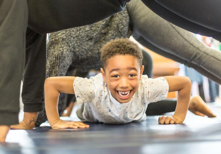 A child smiling and crawling on the ground underneath people who are planking