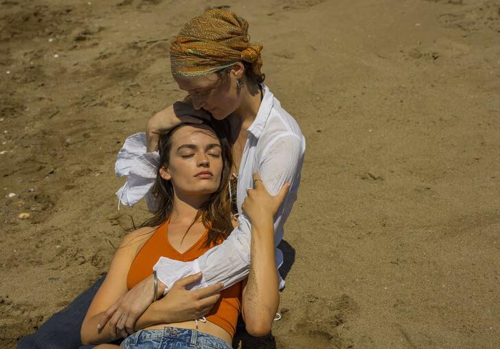 Two women sit on the beach, in each others arms
