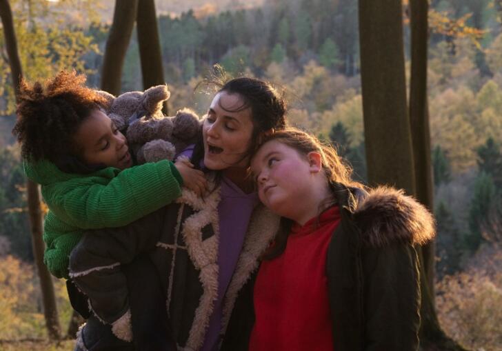 A mother holds her child in a green coat and hugs her elder daughter to her side, in the woods